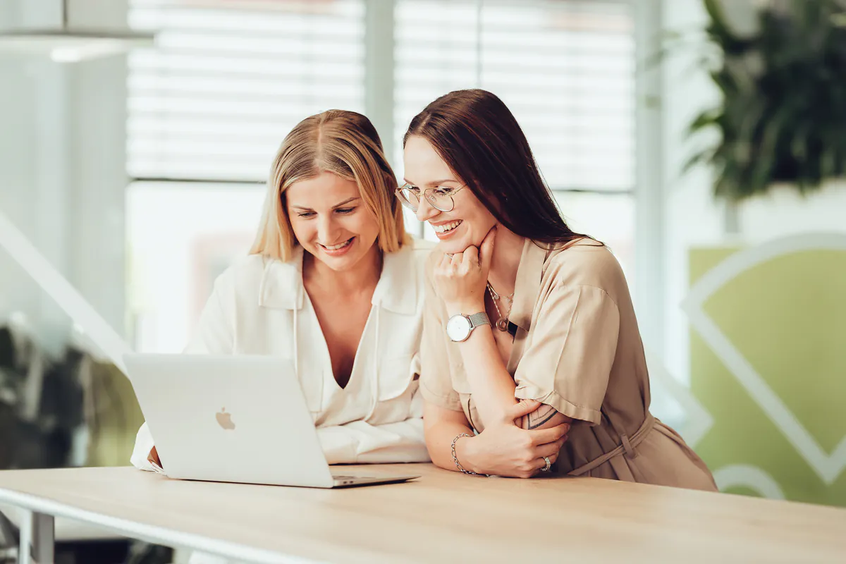 zwei junge Frauen lehnen auf einen Tisch und schauen lächelnd in ein Macbook